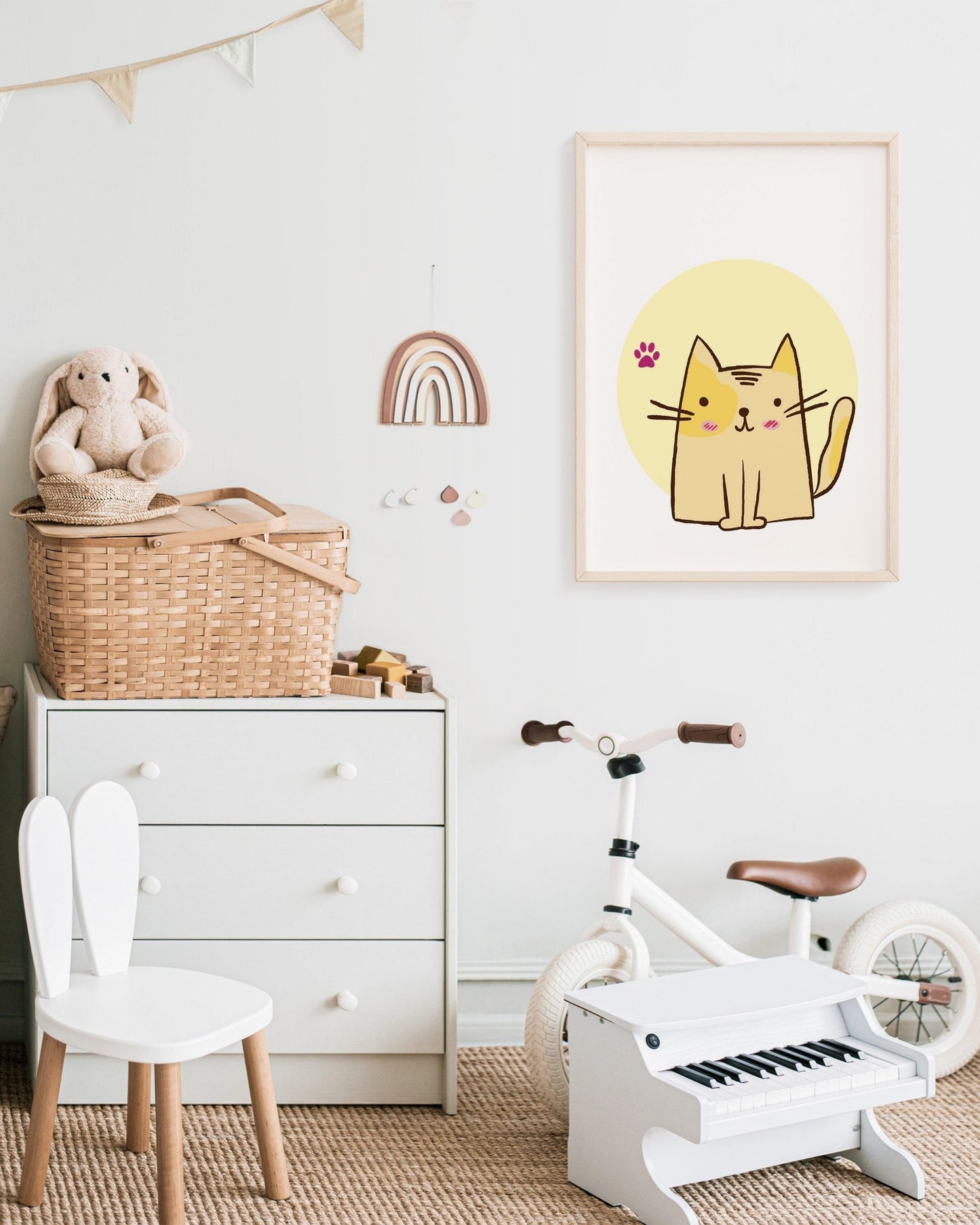 Poster con gattino stilizzato e zampina rosa appeso in cameretta luminosa con cassetto bianco, bici, pianoforte giocattolo e decorazioni arcobaleno.