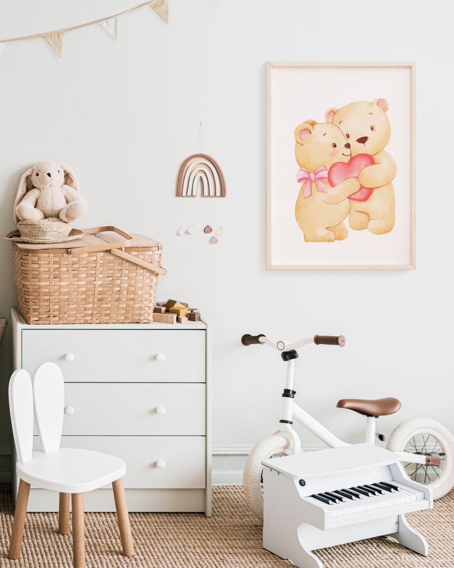 Poster decorativo con due orsetti che si abbracciano tenendo un cuore rosa, appeso sopra cassettiera bianca con giochi e bici per bambini.