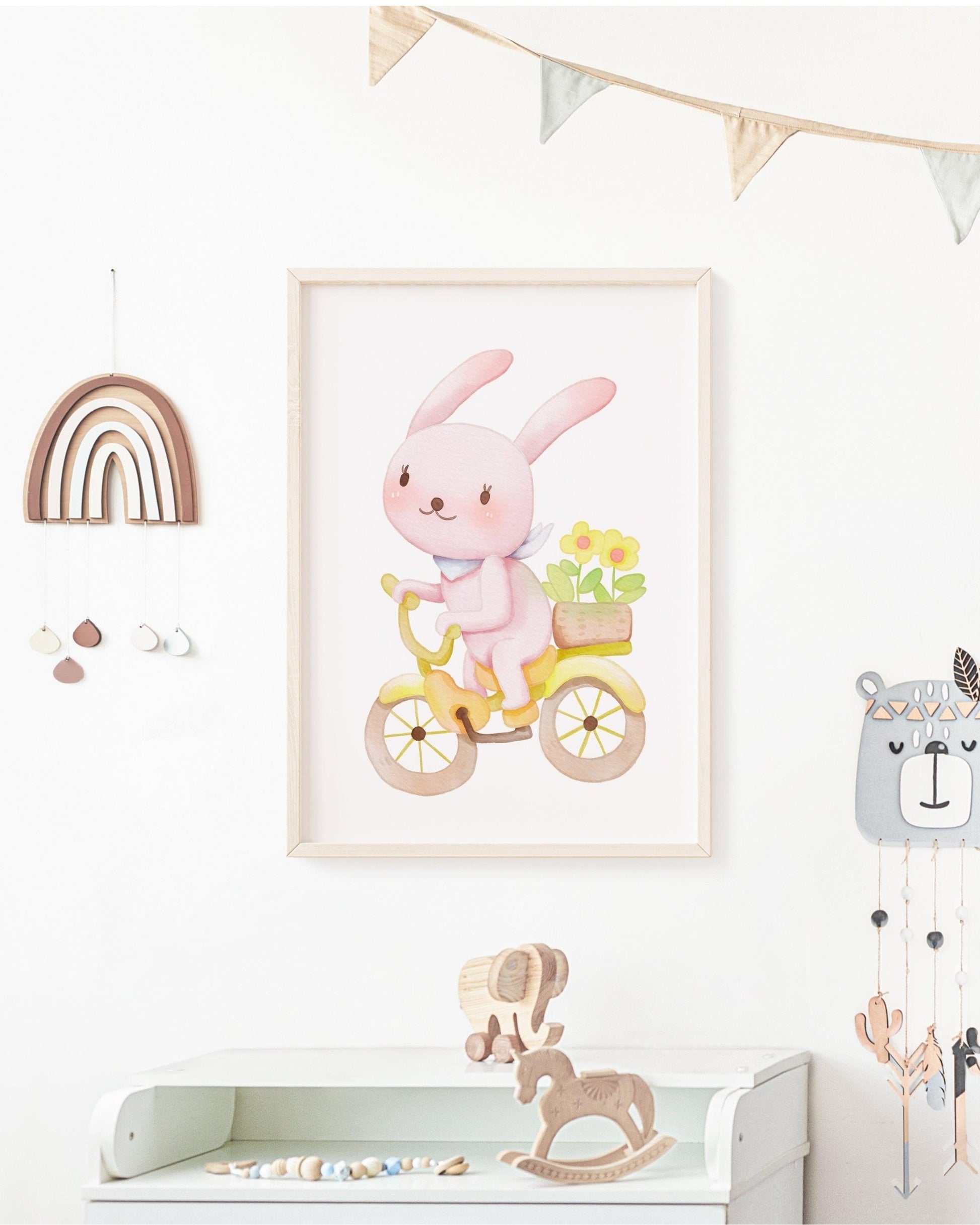 Poster con coniglietto rosa in bicicletta appeso alla parete di una cameretta, con decorazioni in legno, arcobaleno e giochi per bambini.
