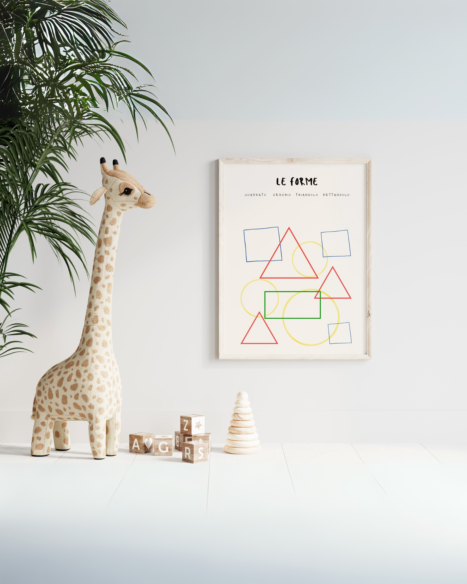 Poster con forme geometriche colorate in cornice chiara, appeso al muro accanto a un peluche a forma di giraffa e giochi in legno sul pavimento.